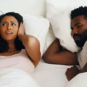 Woman covering ears, man sleeping, snoring issue.